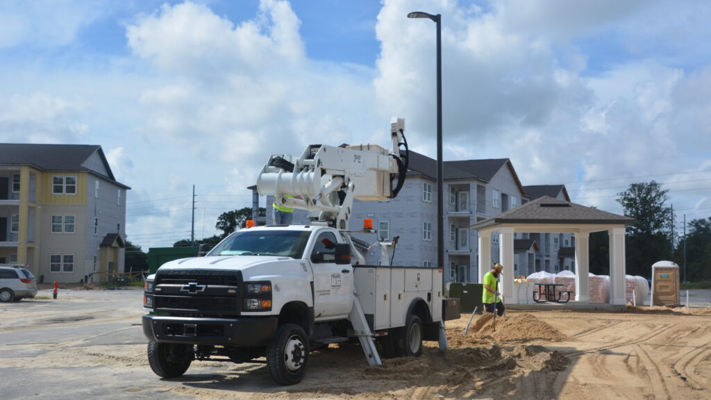 picture of bucket truck with the bucket down. there are apartments in the background mid construction. behind the truck to the right is an electrician in a neon green shirt next to a standing light pole. sky is blue and bloudy.