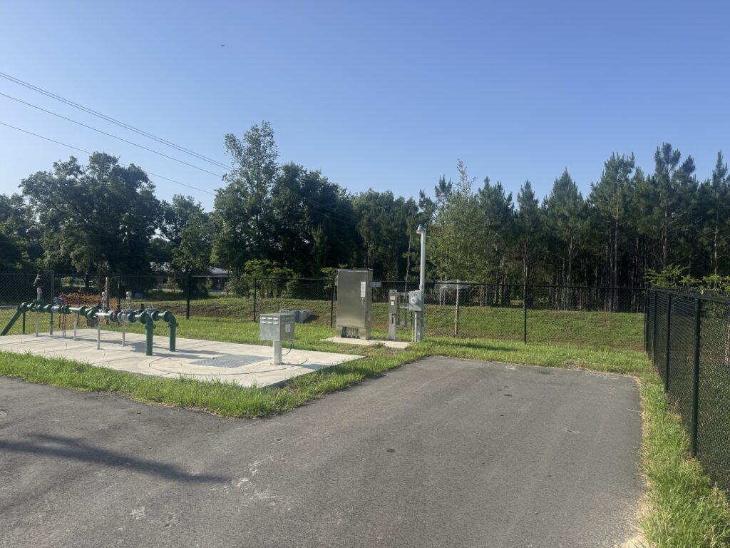 picture of a lift station which includes alumnum or steel metal panels. there is a fence behind the lift station and trees. the sky is blue and clear of clouds.