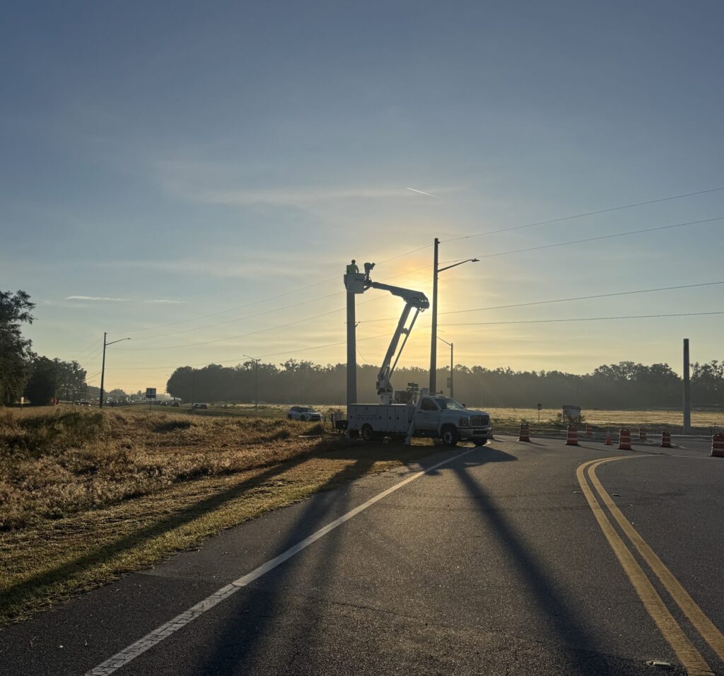 Electrician up in a bucket truck around sunrise with the sun just hitting the large arm holing up the bucket. electrician is near the top on a concrete pole used for holding traffic signal cable.