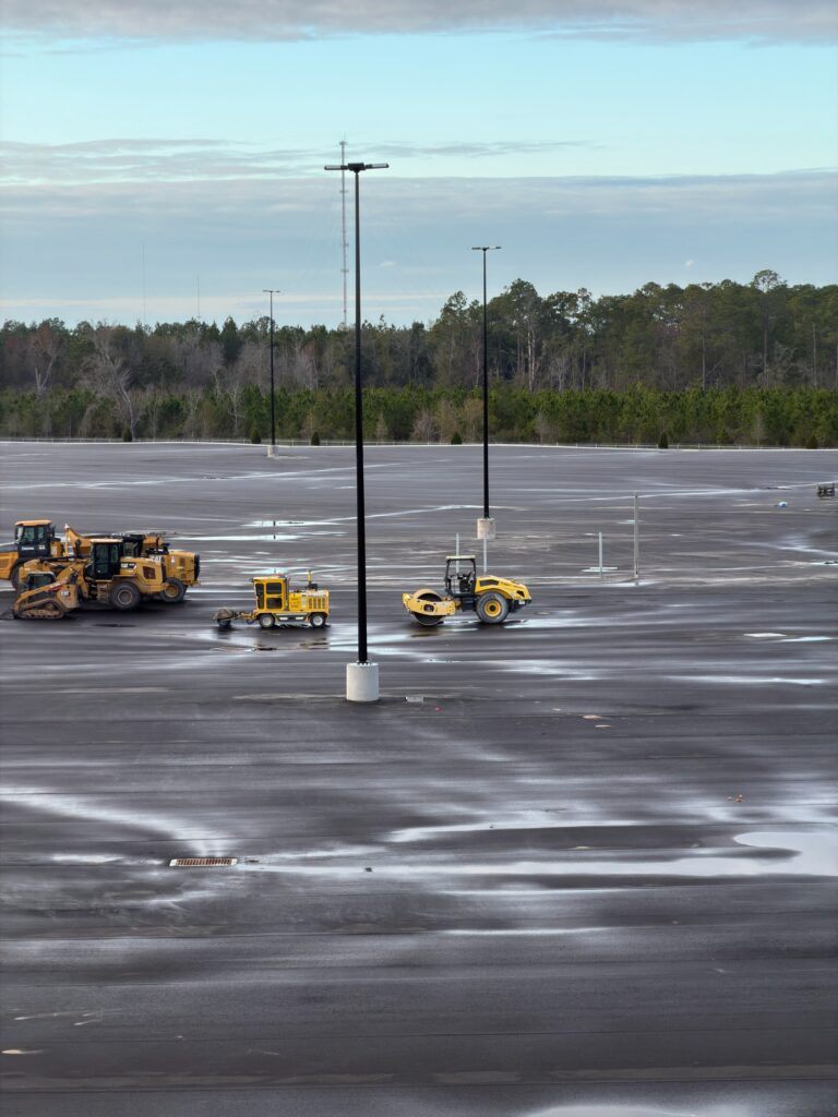 far away picture of site lighting poles which are extremely tall. on the ground next to the poles are large pieces of operating equipment used for rolling out asphault.