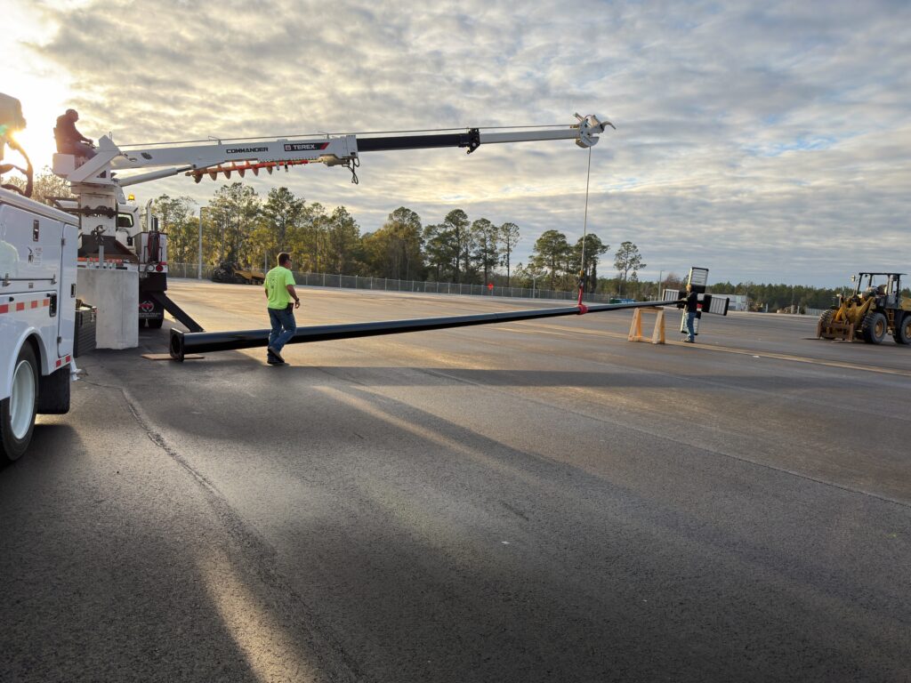 Picture of one worker controlling the line truck that is attached to a light pole lifted slightly off the ground. Another electrical worker wearing a neon green shirt is standing on the ground ready to direct the pole where it needs to go.