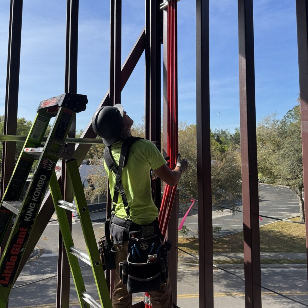 Electrician wearing a neon green shirt and a black hardhat is holding up red conduit in an open building which is mid construction. Trees can be spotted in the background and a short green ladder is right next to the electrician